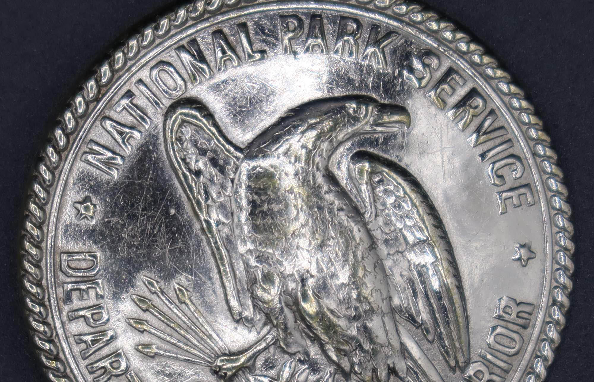 Round silver badge with an eagle in the middle center, marked National Park Service, Department of the Interior. 